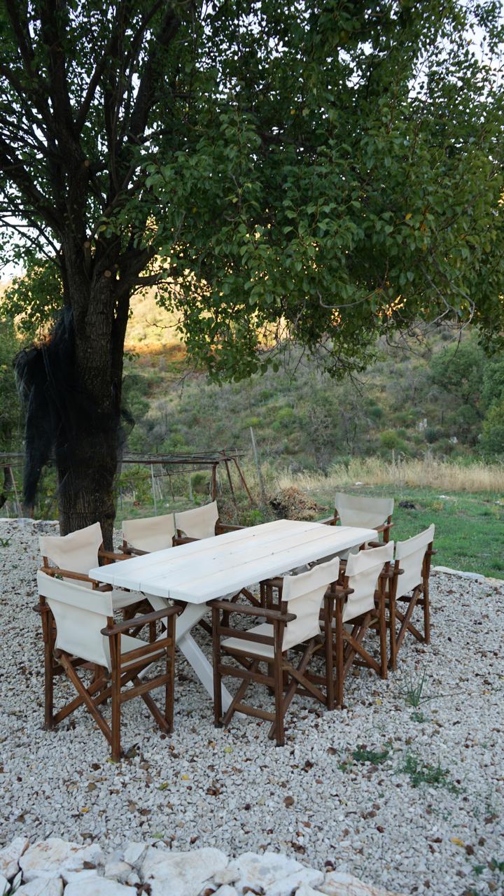 The dining table and canvas chairs set on the white pebble circle under the tree, evening light on the hillside