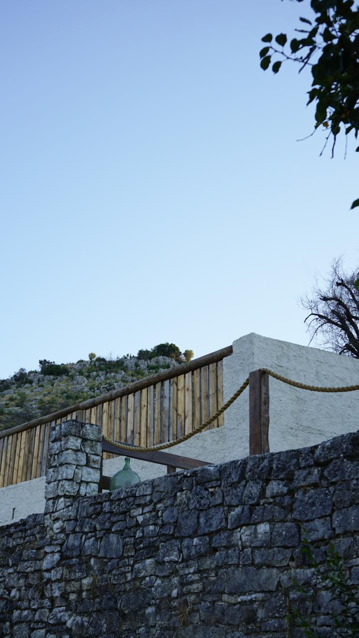 Looking up at the terrace from below the dry-stone wall, a glass demijohn resting along the timber railing
